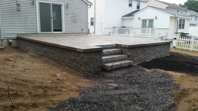 A newly constructed stone patio with steps leading down to a dirt yard, attached to a house with white siding and large sliding glass door.