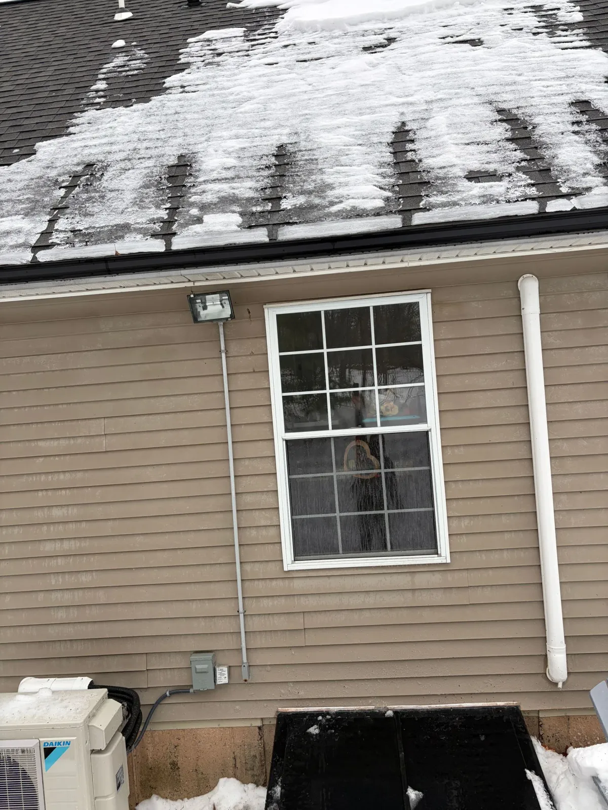 House exterior with tan siding, a white-framed window, and snow on the roof showing dark cleared patches.
