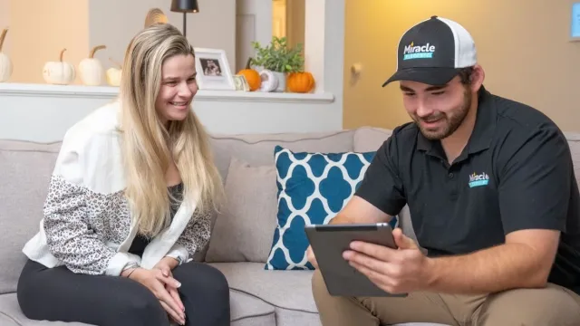 A woman smiling and sitting on a light-colored couch while talking to a man who is showing her something on a tablet. The man is wearing a black polo shirt and a baseball cap with the logo 'Miracle'. There are decorative pumpkins, potted plants, and picture frames in the background.