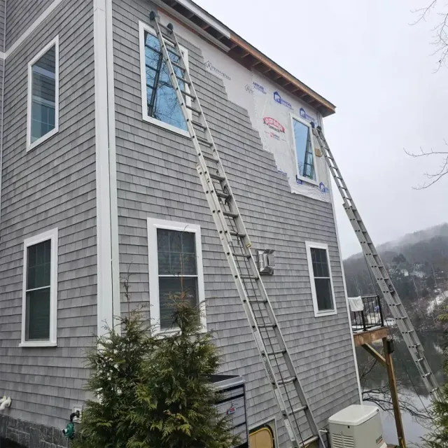 Two extension ladders lean against the gray-shingled exterior of a two-story house undergoing siding repair.