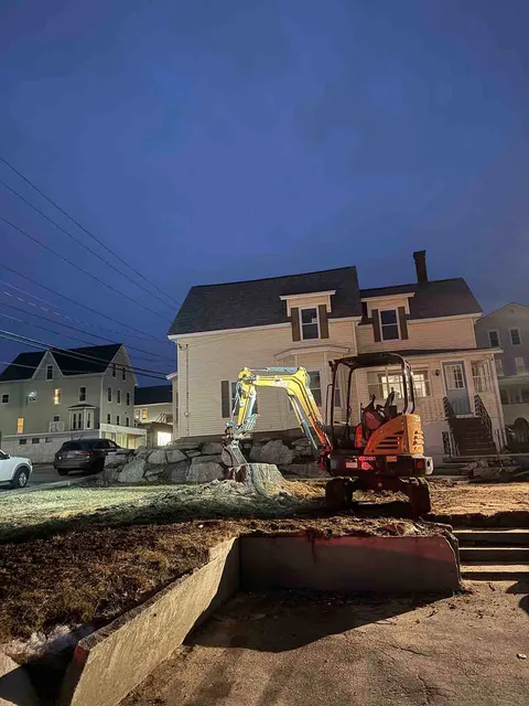 Excavator working in front of a house at dusk, with a bulldozer blade and debris in the yard.