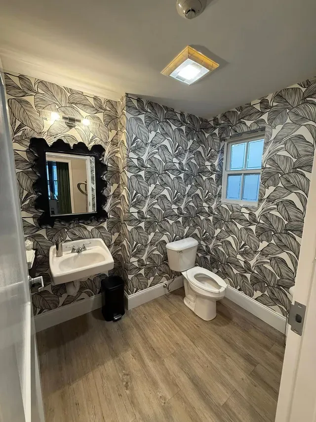 Small bathroom with patterned wallpaper, sink and mirror on left, toilet by window, and wood-look floor.
