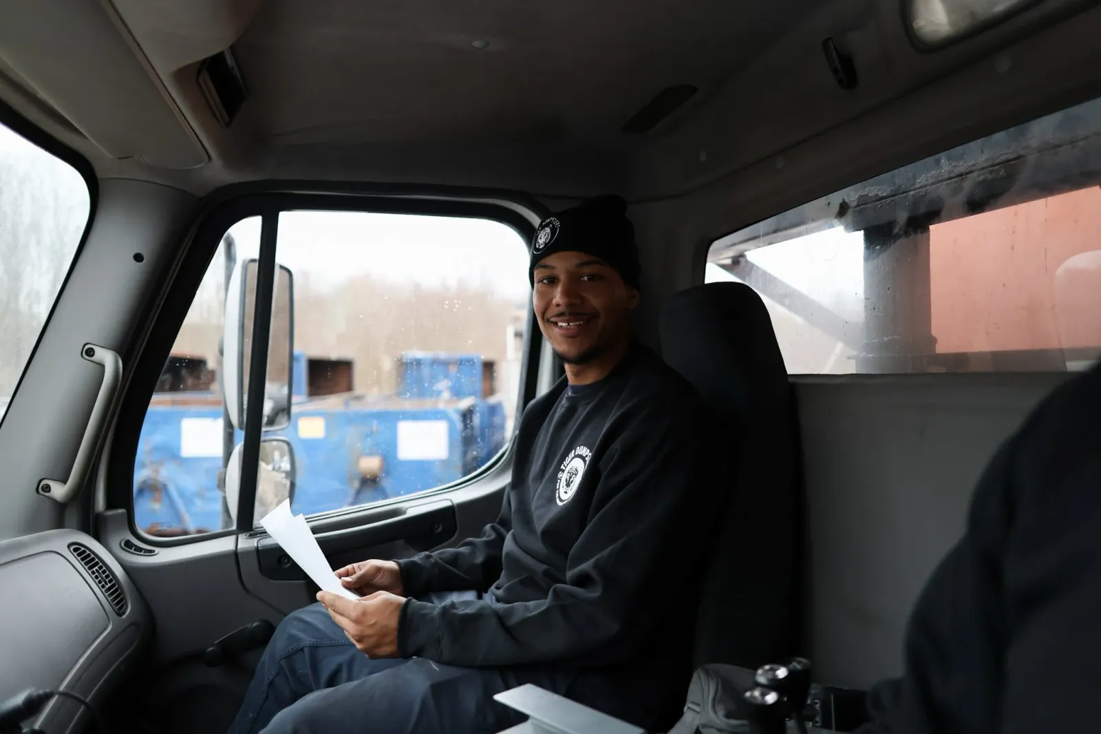 A young man sitting inside a truck cabin, smiling, wearing a black beanie and sweatshirt, holding a piece of paper, with a view of a cloudy outdoor scene through the window.