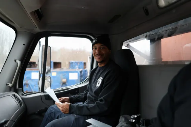 A young man sitting inside a truck cabin, smiling, wearing a black beanie and sweatshirt, holding a piece of paper, with a view of a cloudy outdoor scene through the window.
