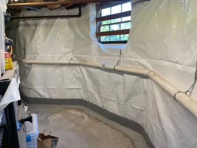 Plastic-lined basement with a curved white pipe, small window, and construction materials on the floor