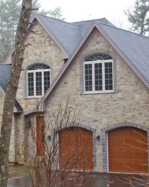 Front view of a two-story house with stone exterior, steep gabled roof, and wooden garage doors. A tree with bare branches partially obscures the house.