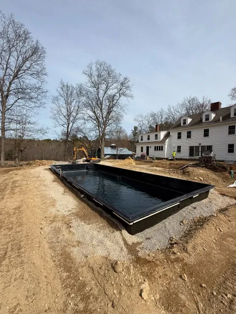 Rectangular black swimming pool shell in a dirt lot, near a white building with red chimneys.