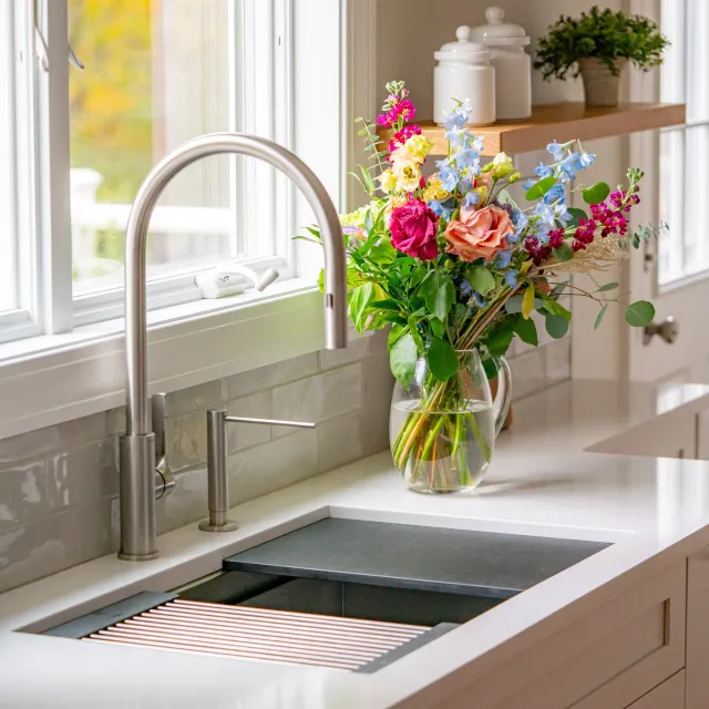 A kitchen sink with a chrome faucet and a vase of colorful flowers on the white countertop near a window.