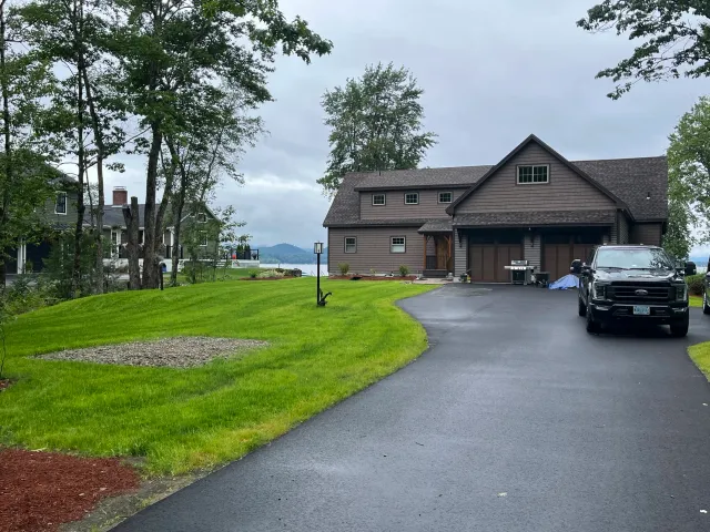 A driveway leading to a dark brown house with a two-car garage, surrounded by green grass and trees, with a black truck parked on the right side and an overcast sky.