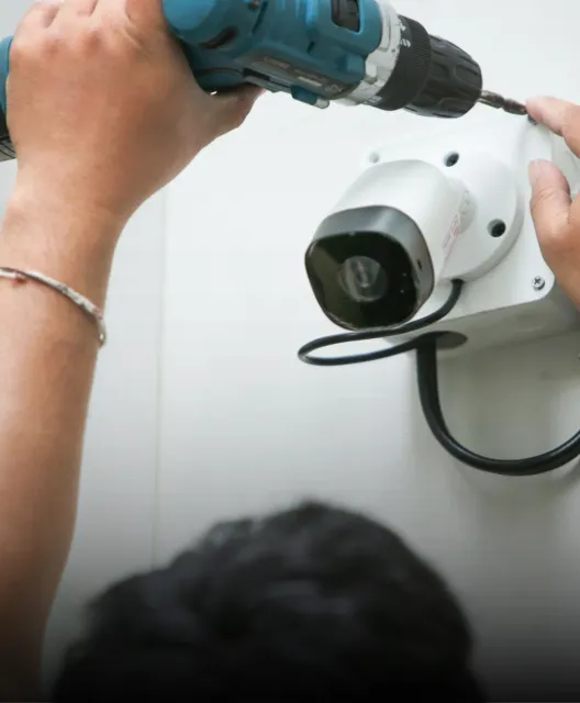 A technician drills into a white wall to mount a security surveillance camera.