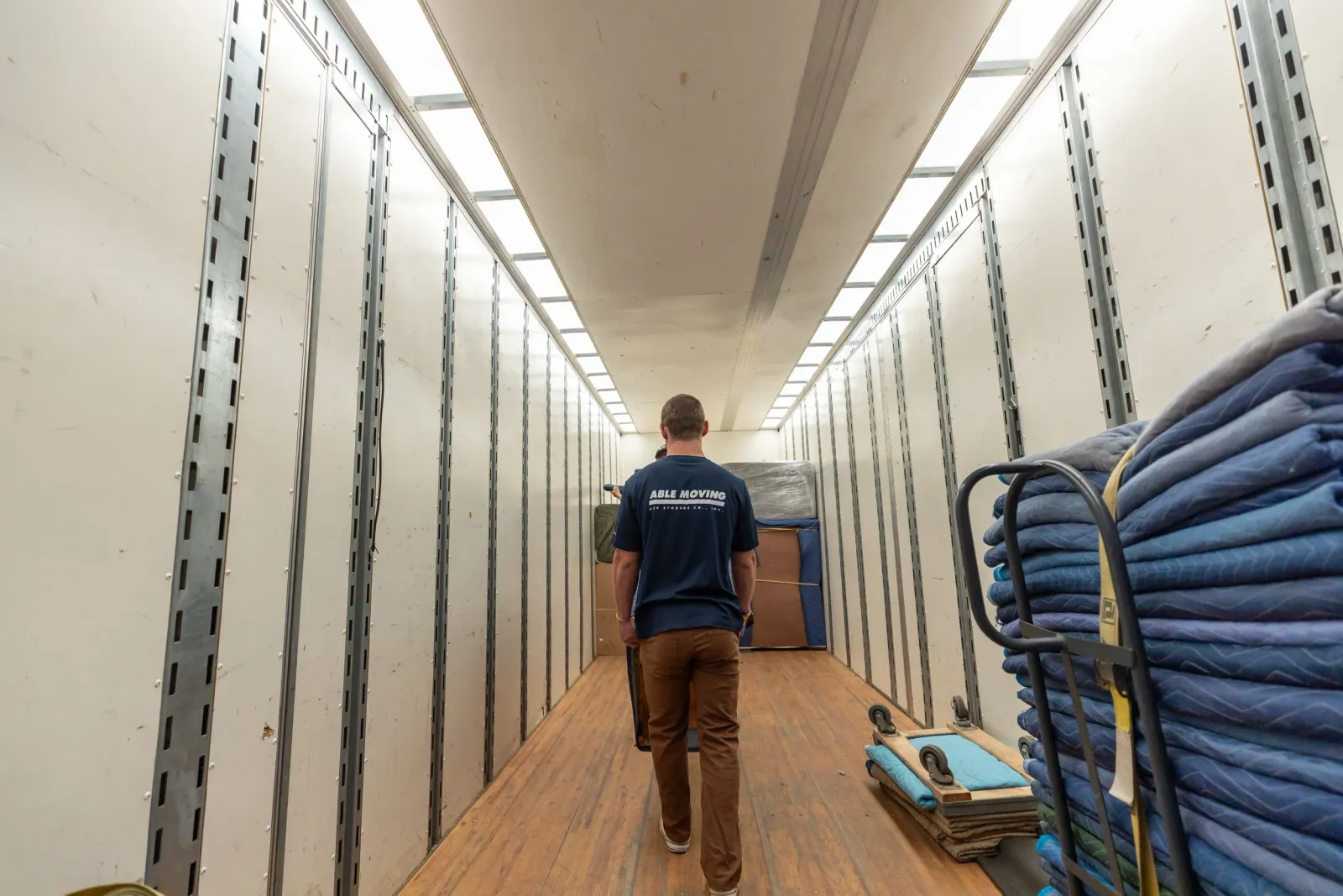 One man walking inside a narrow box truck with metal walls, hardwood flooring, and stacked blankets on a dolly on the right side.