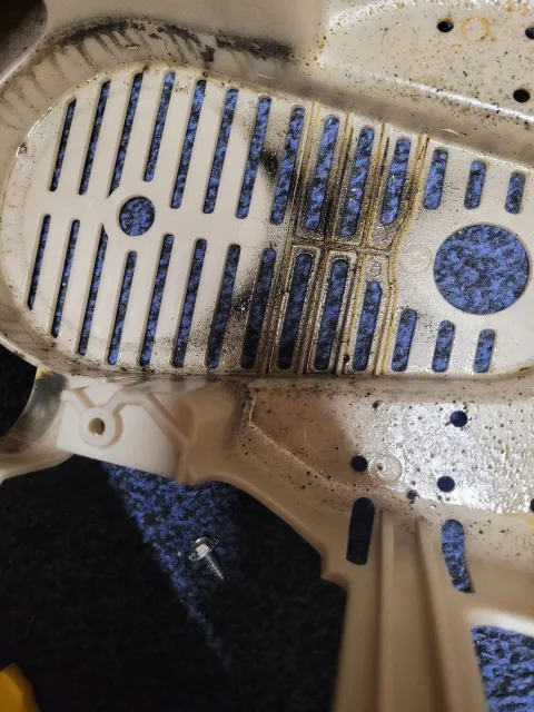 A close-up of a dirty, beige plastic machine casing featuring a slotted intake vent covered in dark dust and debris.