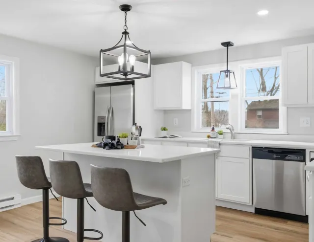 Modern kitchen with white cabinets, island with three gray stools, stainless steel fridge and dishwasher.