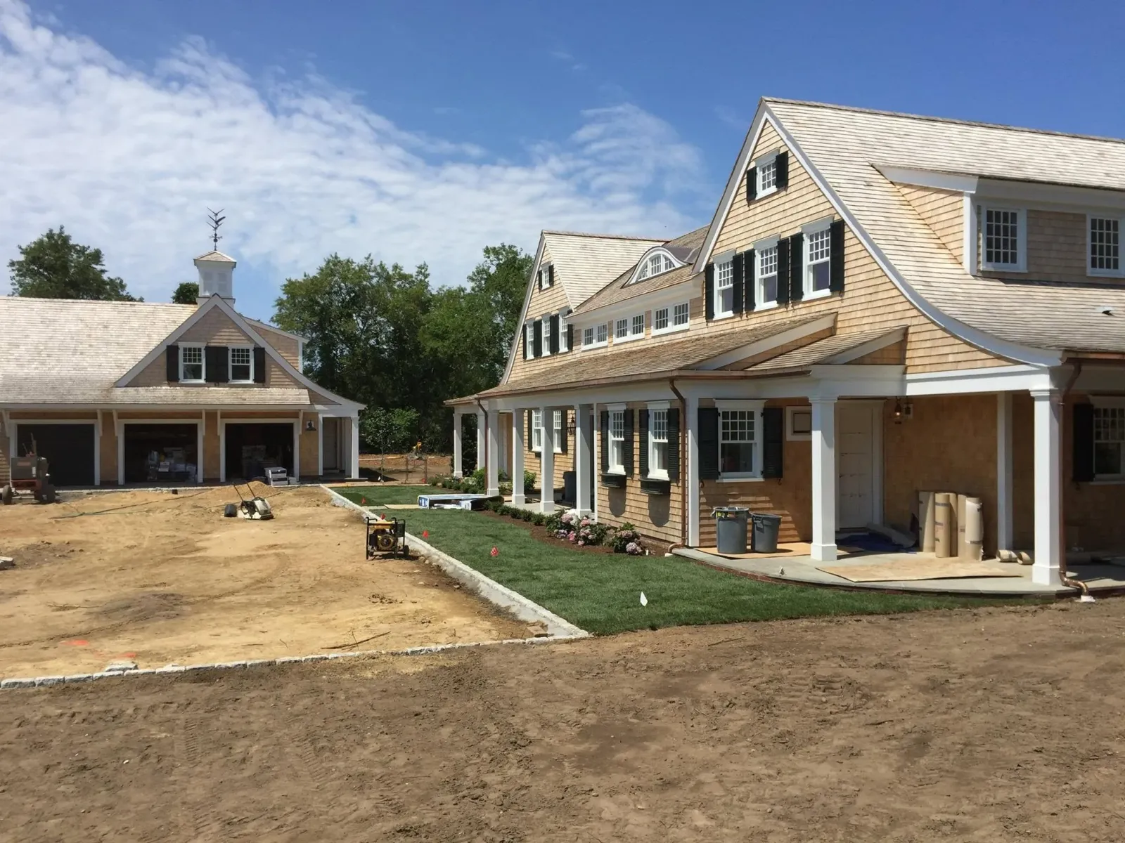 Residential construction site with a house featuring beige shingles, black shutters, white trim, and a porch. The yard has been recently landscaped with a green lawn and flowers. An adjacent garage is under construction, and construction equipment is present on the dirt ground. The sky is partly cloudy.
