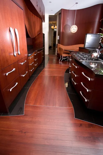 Interior of a kitchen with polished dark wood cabinetry, black countertops, and a wooden floor with a unique curved design; visible in the background are chairs, a pendant light, and a doorway.