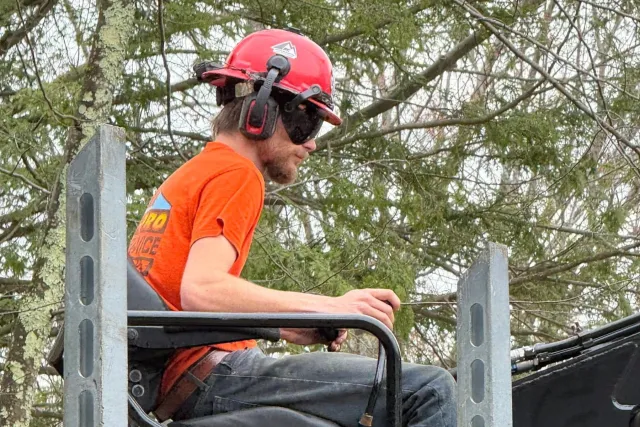 A man in construction gear, including a red helmet and orange shirt, working on a utility pole with trees in the background.