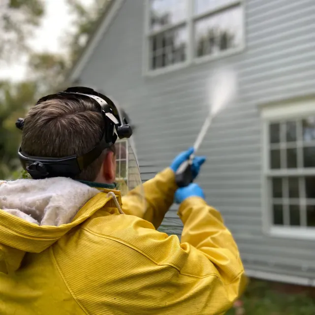 Worker in a yellow rain jacket and face shield washing gray clapboard siding with a pressure wand