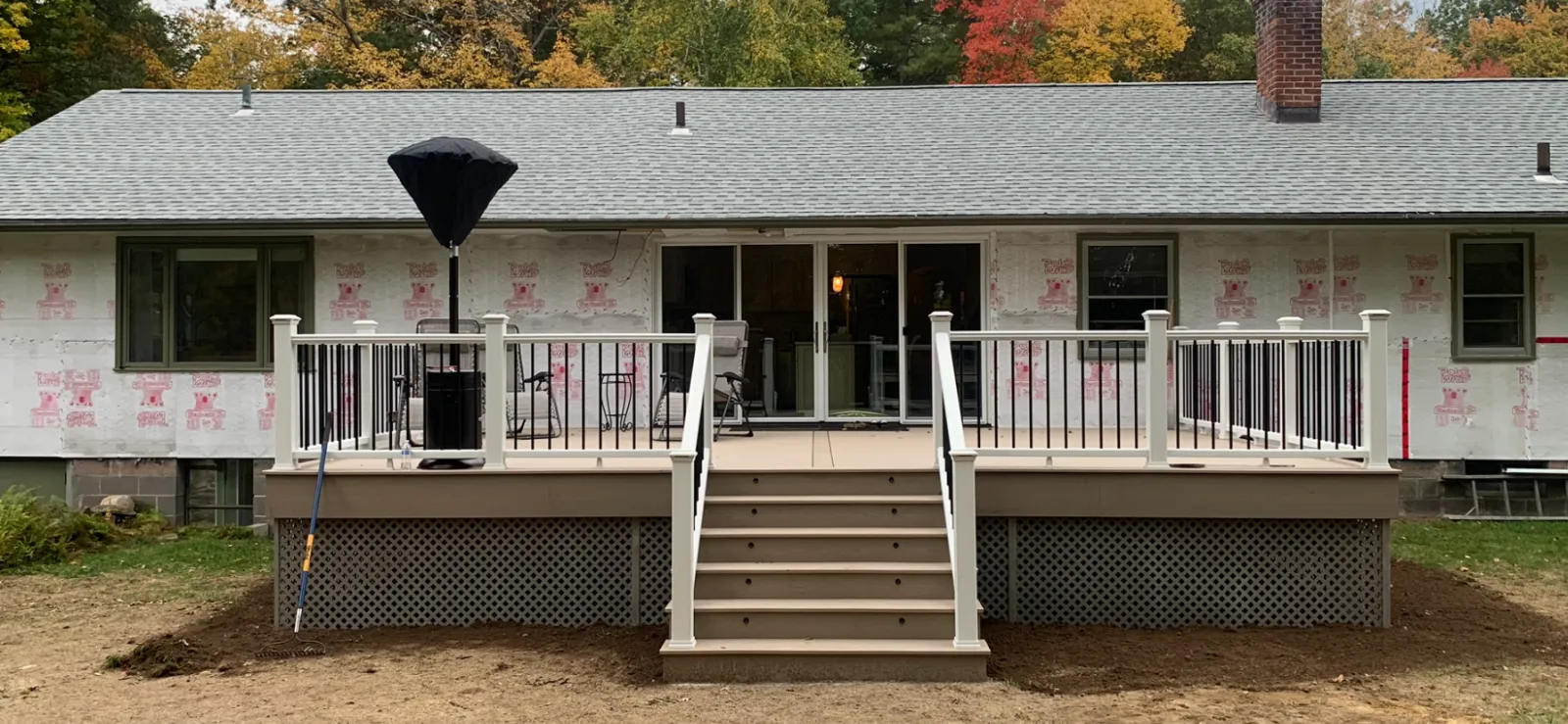 Backyard deck under construction on a house with a gray roof, black and white railing, steps leading down to a dirt yard, and a large patio umbrella.
