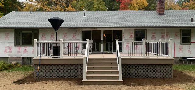 Backyard deck under construction on a house with a gray roof, black and white railing, steps leading down to a dirt yard, and a large patio umbrella.