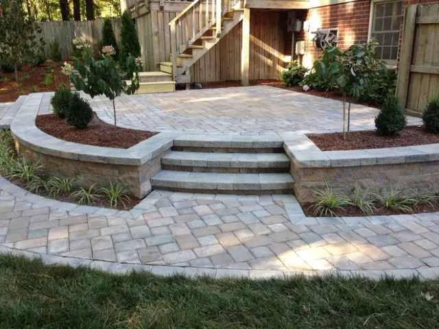 Stone patio with curved steps, paver walkway, and raised garden beds in a backyard