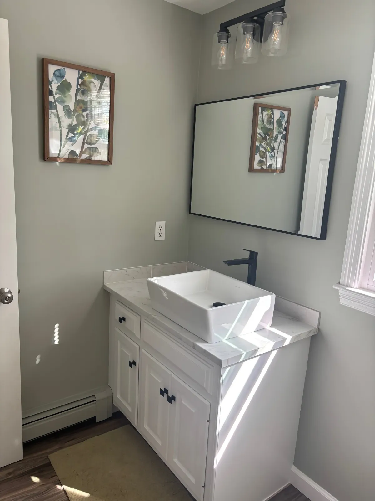 A bathroom with light green walls, a white vanity with black knobs, a vessel sink, a rectangular mirror, a black faucet, a framed botanical print, and a three-light vanity fixture.