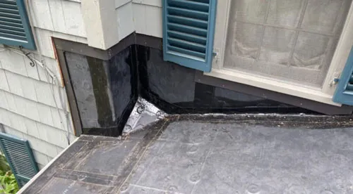 Chris Battaini Roofing - A flat rooftop covered with black rubber material. Theres a corner where the roof meets the house, with siding, a window, and blue wooden shutters.