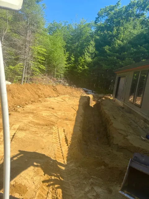 Construction site with a cleared and dug-out area next to a house, surrounded by trees under a clear blue sky.