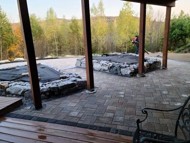 A person uses a leaf blower on a brick patio under construction, with tiered stone planters and a forest in the background.