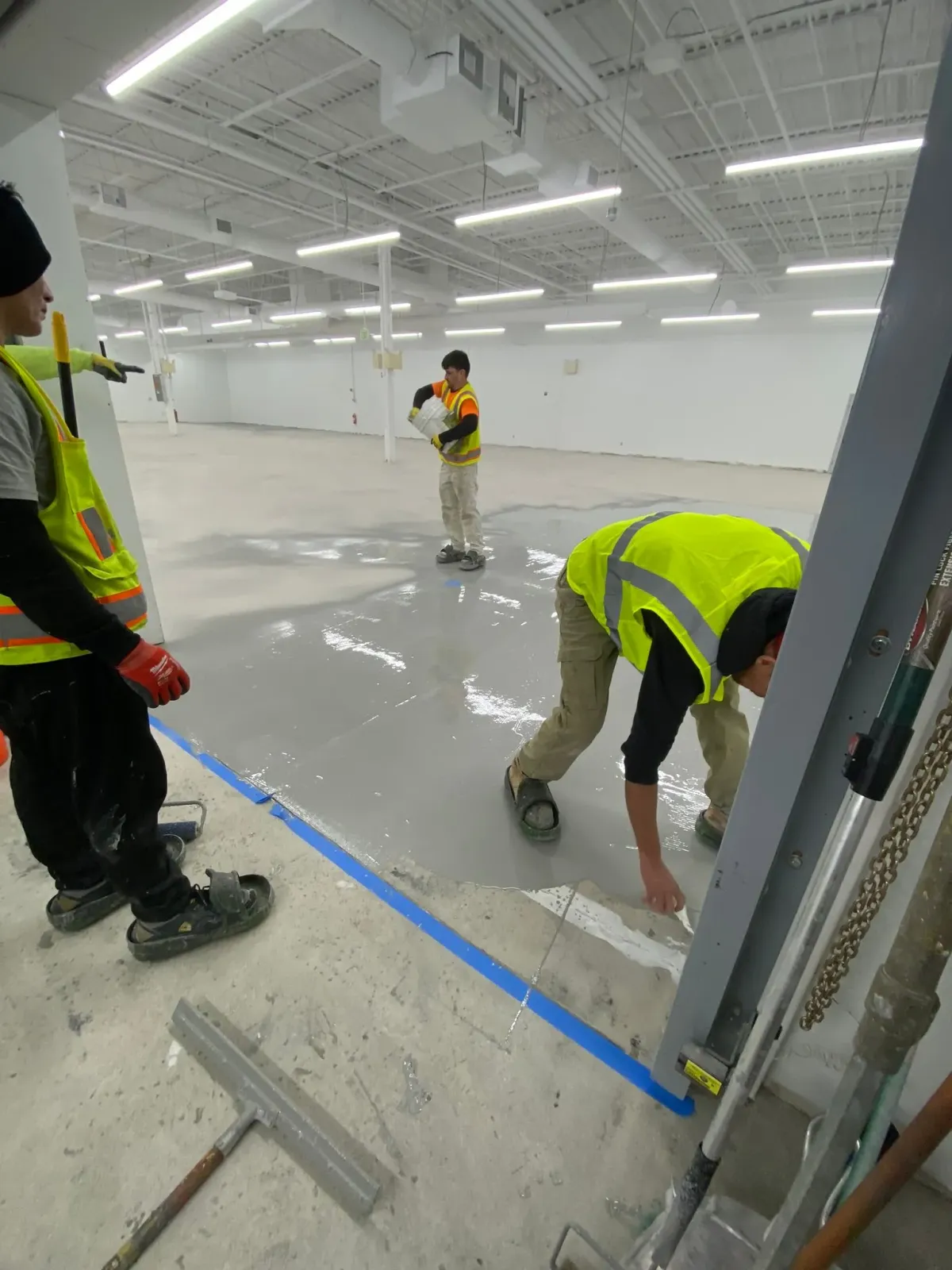 Workers in neon vests applying blue tape on a shiny warehouse floor under bright lights
