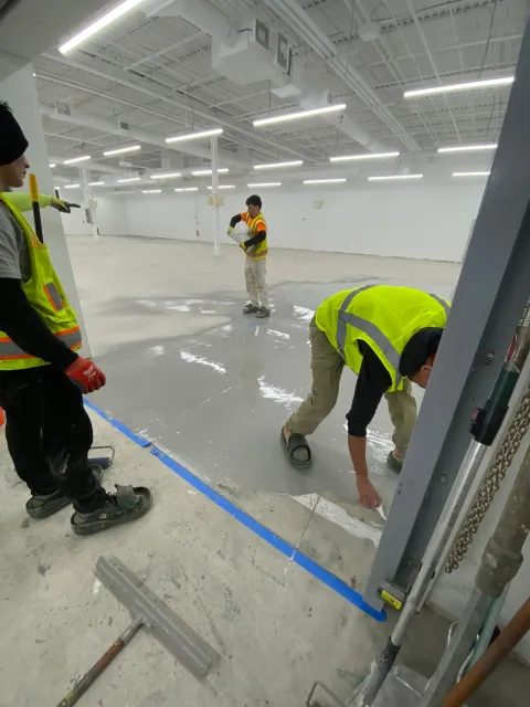 Workers in neon vests applying blue tape on a shiny warehouse floor under bright lights