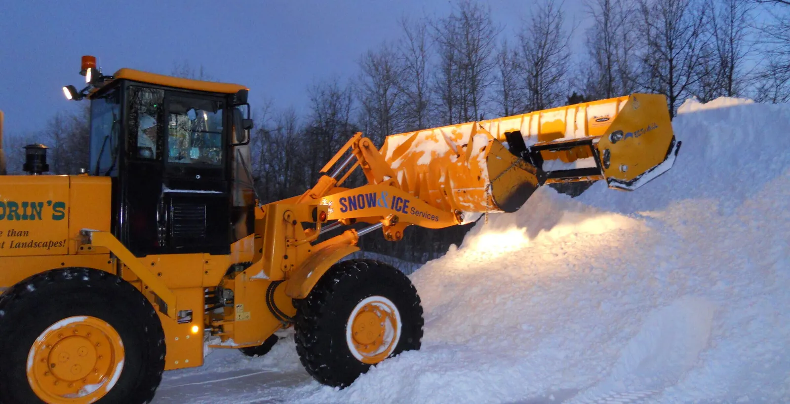 A yellow snowplow clears a large pile of snow in a parking area, surrounded by bare trees under a cloudy winter sky.