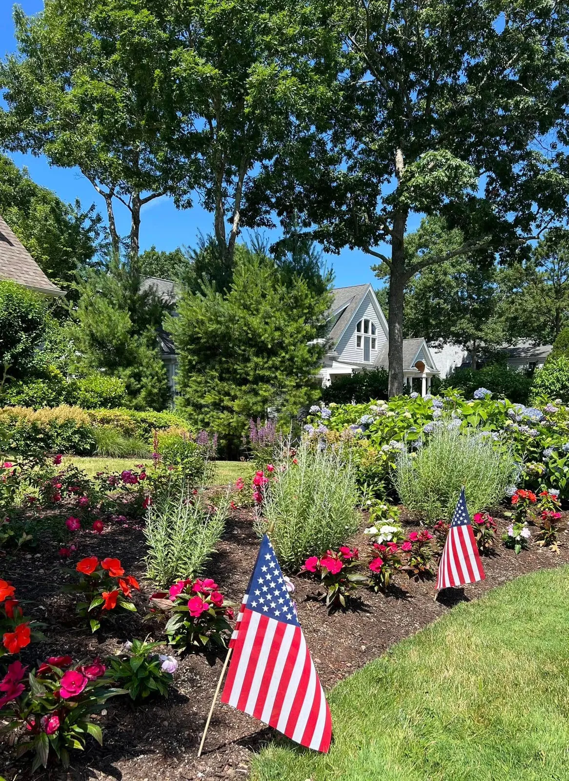 American flags in a flower garden with a house in the background. Green trees and blue sky.