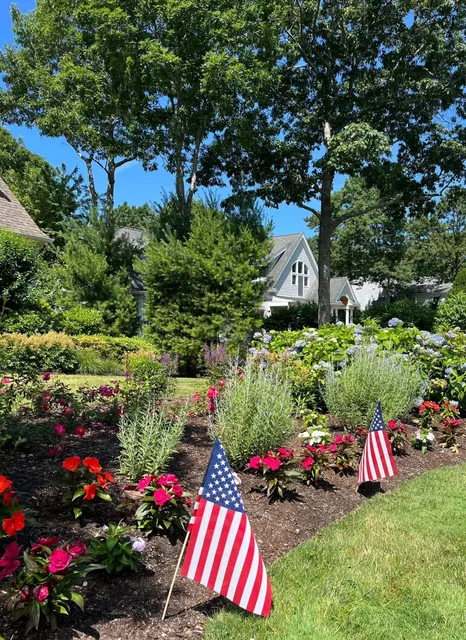 American flags in a flower garden with a house in the background. Green trees and blue sky.