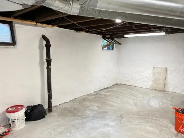 Basement interior with white walls, exposed ceiling, concrete floor, and pipes. Buckets and tools are on the floor.