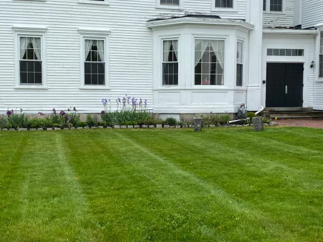 White house with adjoining garage, fronted by a neatly mowed lawn and a flower bed with purple flowers.