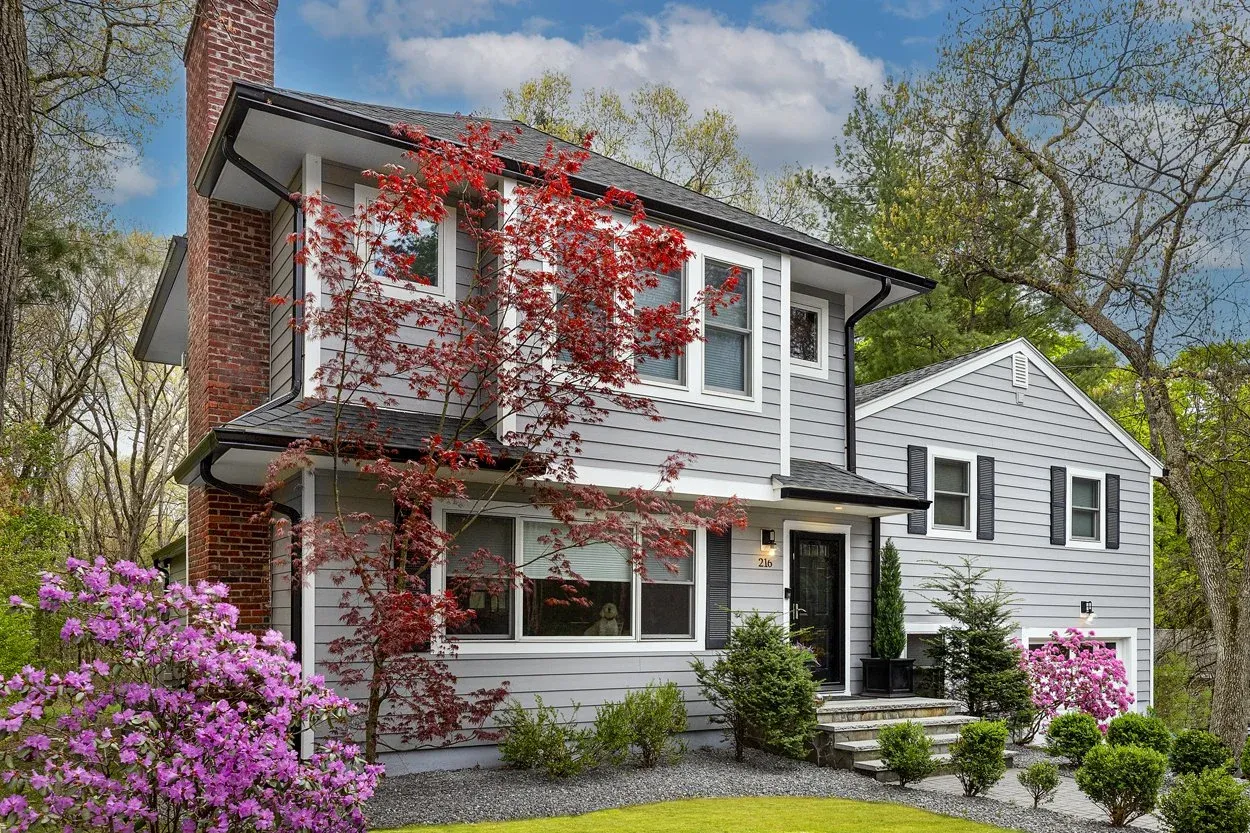 A two-story house with gray siding, black shutters, and a brick chimney, surrounded by flowering bushes and trees, with a well-maintained lawn and stone steps leading to the front door.