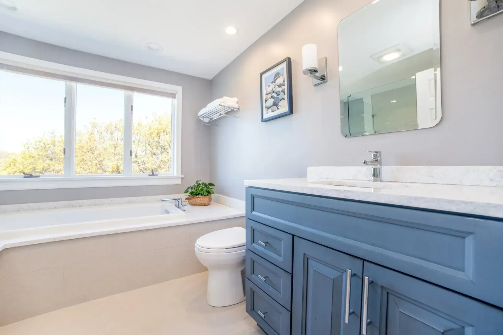 Bright modern bathroom with blue vanity, white tub, and large window.