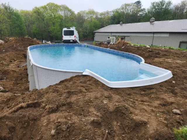 Above-ground rectangular swimming pool with water, partially installed in a backyard with dirt surrounding it, and a white van and building in the background.