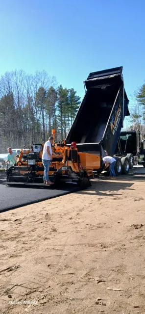 Asphalt paving construction site with a dump truck unloading into a paving machine. Workers are present.