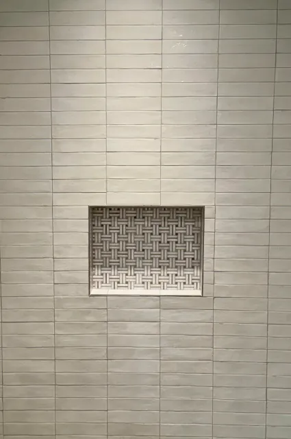 A wall of light beige horizontal rectangular tiles with a small square vent featuring a geometric pattern near the lower middle of the image.