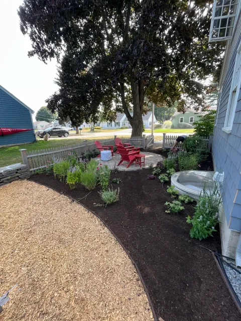 A backyard with a large tree, red chairs on a stone patio, landscaped garden beds with plants and flowers, a small fountain, and a person working in the garden. There is a blue house to the right and a wooden fence around the yard.