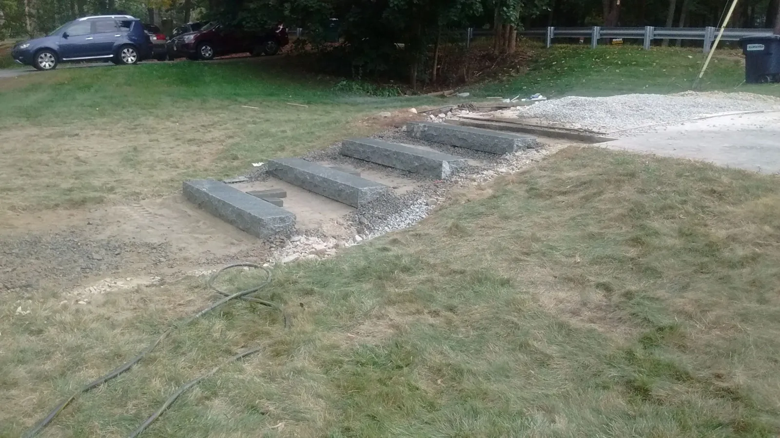Granite steps under construction on a grassy slope, surrounded by loose gravel, dirt, and construction materials, with a guardrail, parked cars, and trees in the background.
