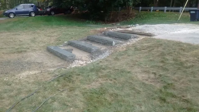 Granite steps under construction on a grassy slope, surrounded by loose gravel, dirt, and construction materials, with a guardrail, parked cars, and trees in the background.