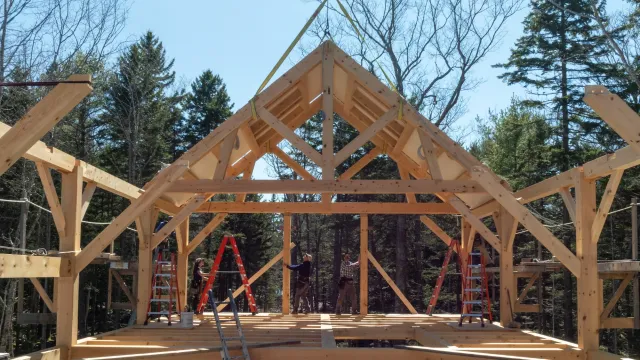 Wooden house structure under construction with workers and ladders, forest in the background.