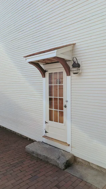 Exterior wall of a building with a white door, small awning, and a wall-mounted lantern, on a brick walkway.