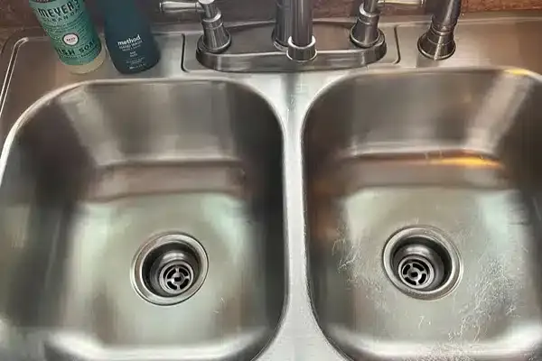 Double stainless steel sink with faucet, soap bottle, and lotion near the edge on the countertop.