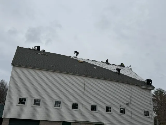 White house with a steep roof and several roofers working on top under a cloudy sky