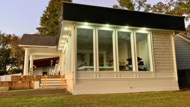 Enclosed sunroom with floor-to-ceiling windows, white trim, recessed soffit LED lighting and adjacent wood deck with illuminated steps — three-season vs four-season rooms in massachusetts