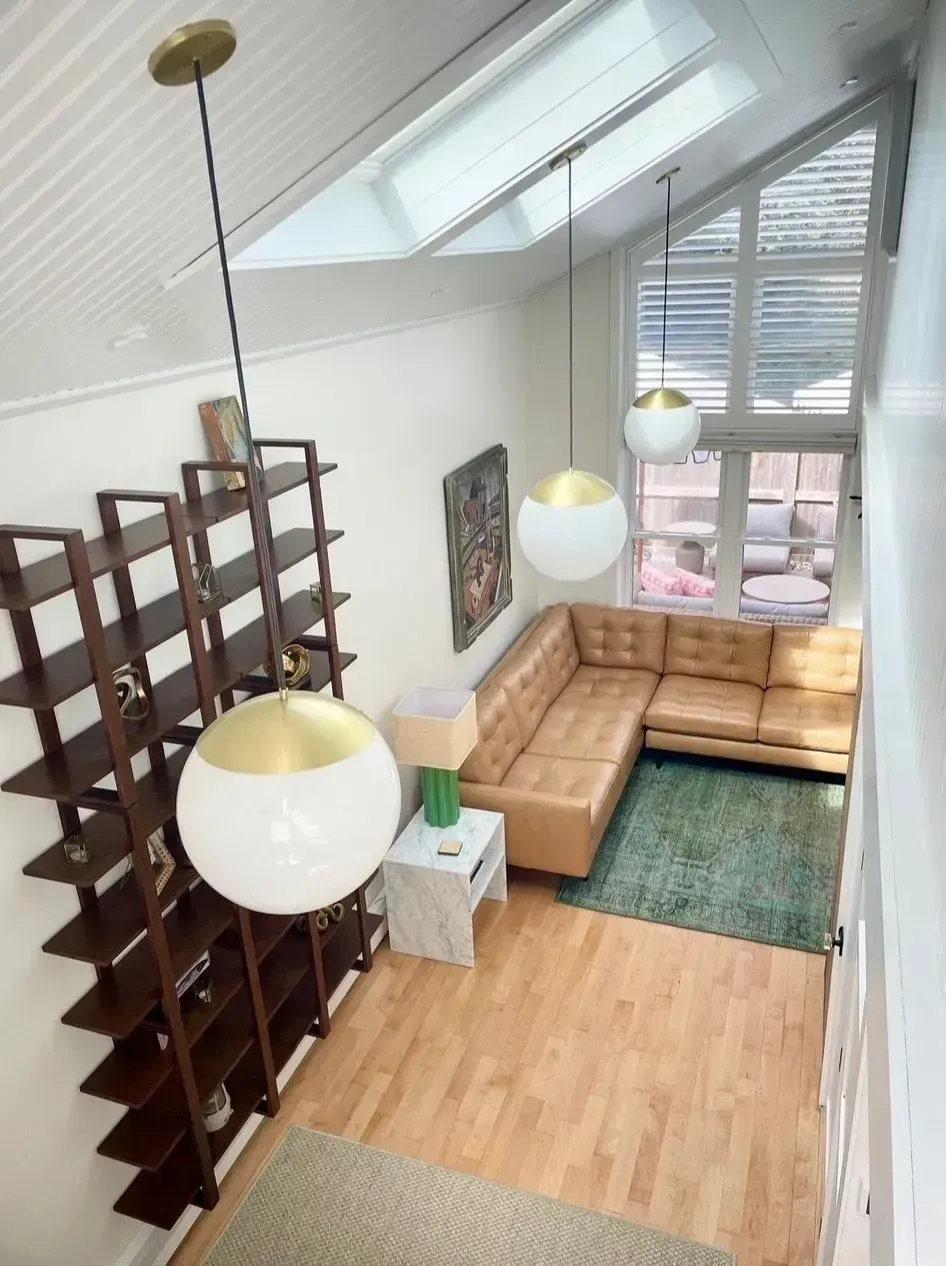 Bright loft living room with tan sectional, green rug, hanging globe lights, skylight, and tall shelving divider