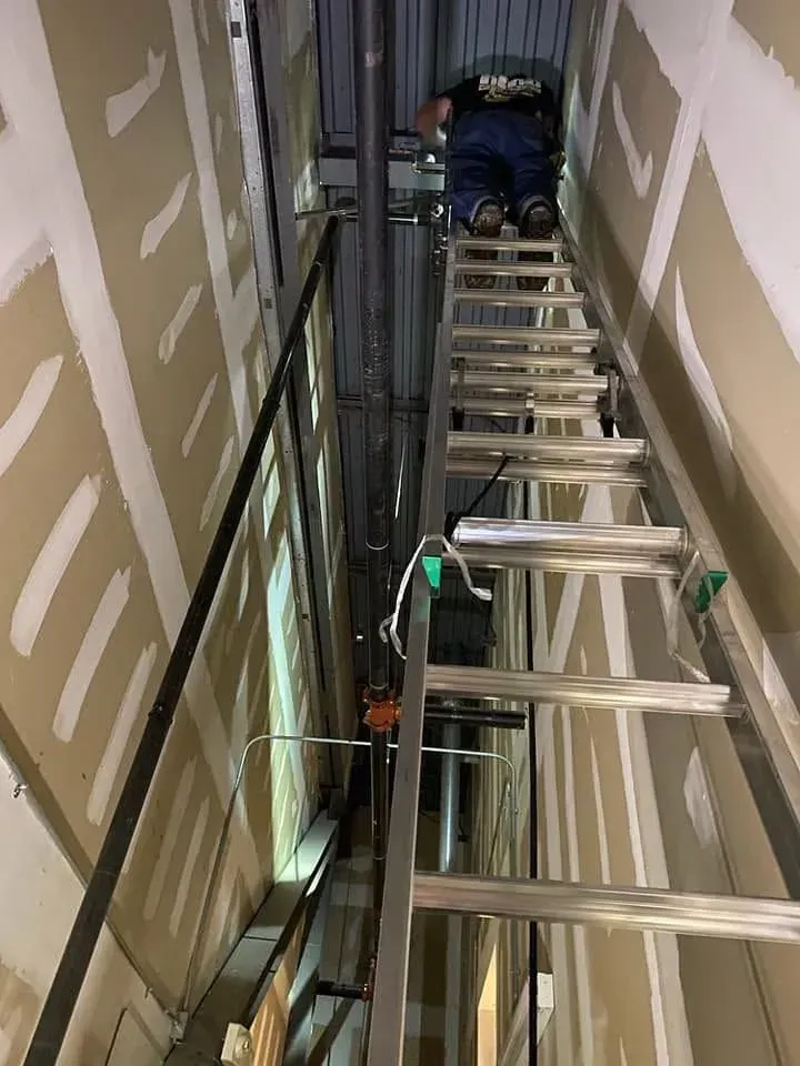 Worker on a ladder in a narrow unfinished hallway near exposed wiring and wall panels